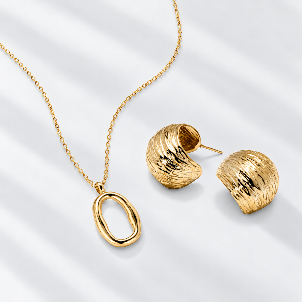 Gold jewelry set featuring a textured oval pendant necklace and matching textured half-ear stud earrings, arranged on a light gray background with soft diagonal shadows.