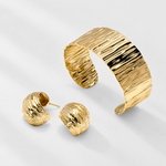 A gold jewellery set featuring a wide, ripple effect textured cuff bracelet and two matching textured stud earrings, displayed on a clean light gray background.