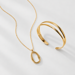 Gold jewelry set featuring a delicate chain necklace with a minimalist oval pendant and a matching double-band cuff bracelet, displayed on a clean white background.