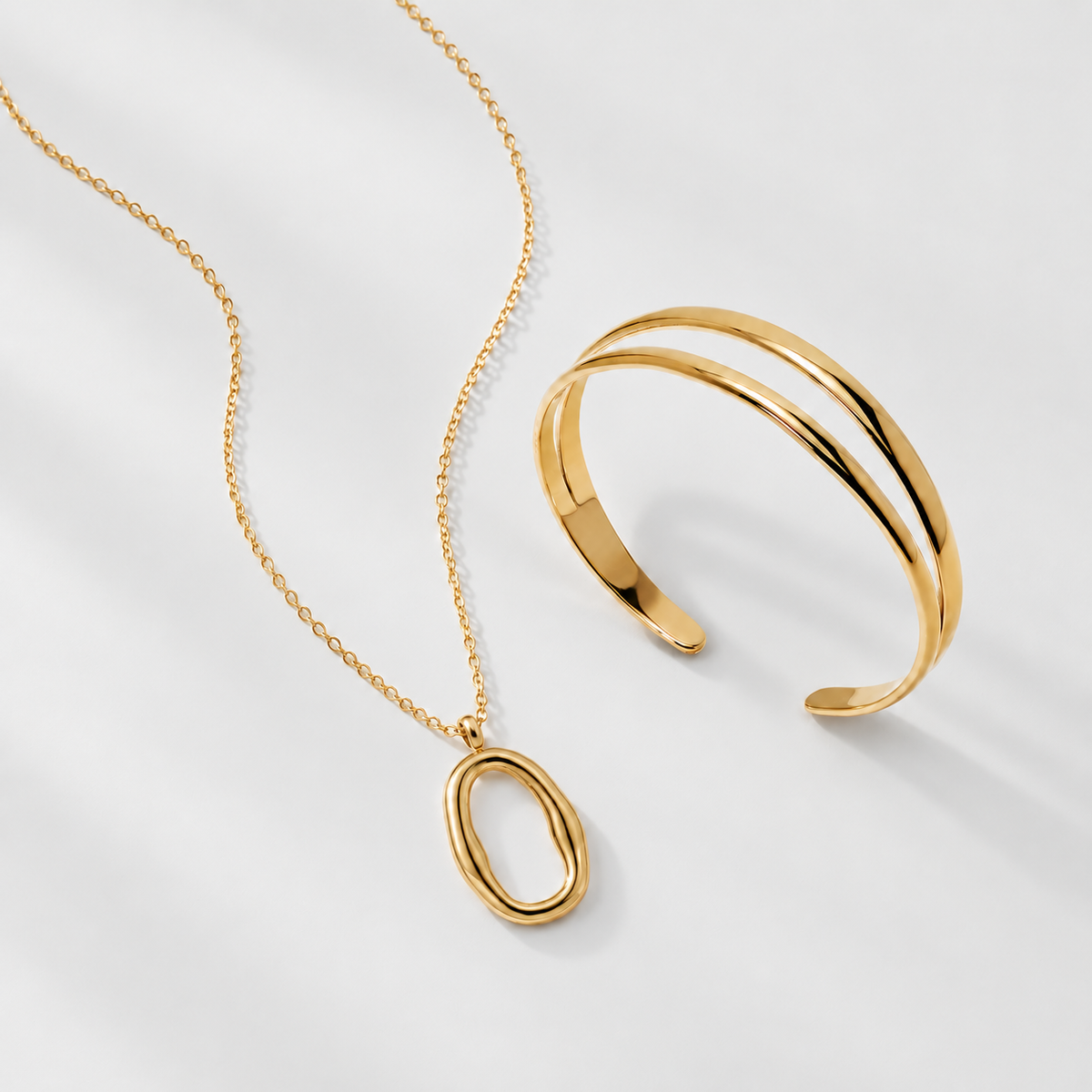 Gold jewelry set featuring a delicate chain necklace with a minimalist oval pendant and a matching double-band cuff bracelet, displayed on a clean white background.