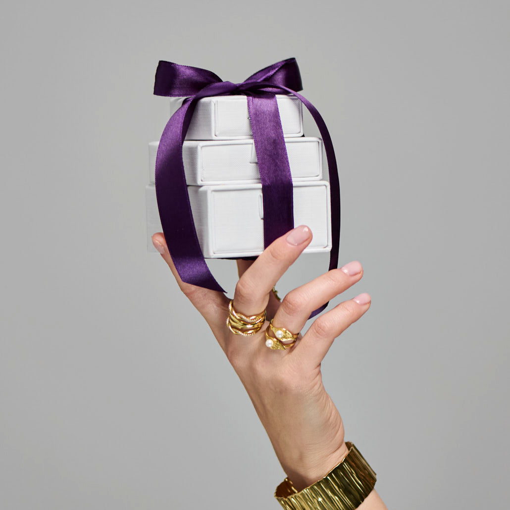 Hand holding a white cylindrical container with a purple ribbon against a gray background